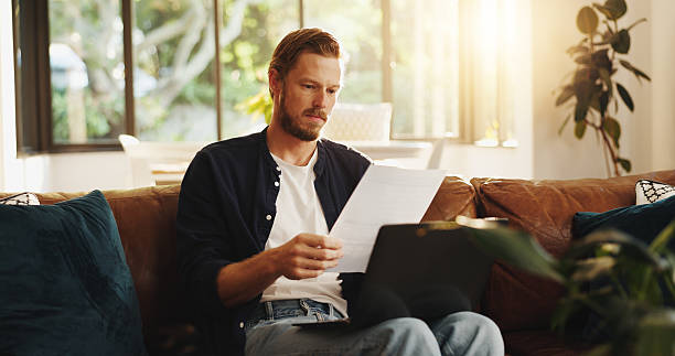 istock Man, paperwork and reading on sofa with laptop, writer and thinking for script draft in home. Person, editor and technology in living room for research, networking and application with freelancer 2225080185