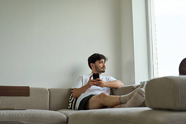 istock Young man relaxing on sofa, holding smartphone and looking out window 2225099856