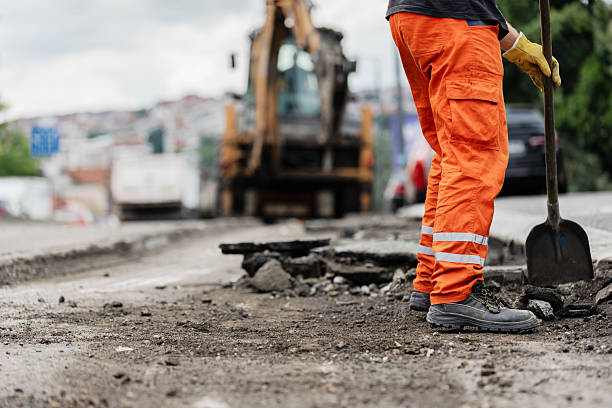 istock Construction worker repairs road surface with shovel while heavy machinery operates in background during cloudy day 2225102245