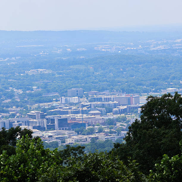 A view from a mountain looking down over Huntsville Alabama on a bright summer day.  Mountains in the distance beyond Huntsville are  covered in haze. Landscape photo is framed with green foliage in the foreground.