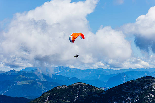 A person glides effortlessly through the sky, suspended beneath a vibrant paraglider, surrounded by stunning mountain ranges and fluffy clouds in a clear blue atmosphere.