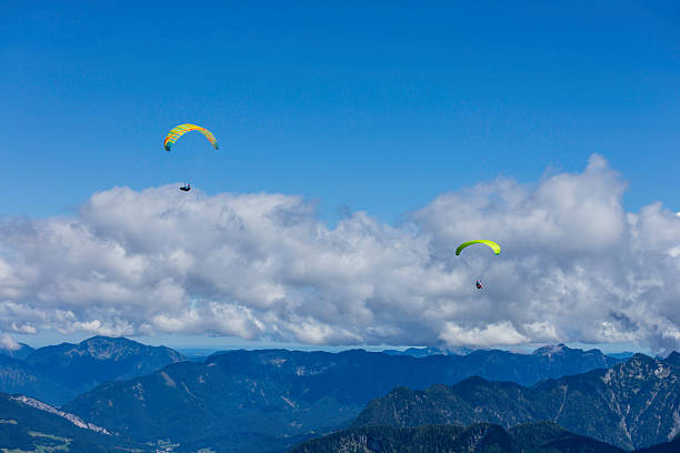 Two individuals glide gracefully in vibrant paragliders high above rugged mountain peaks, enjoying the thrill of the sport during a clear day in the Alps.