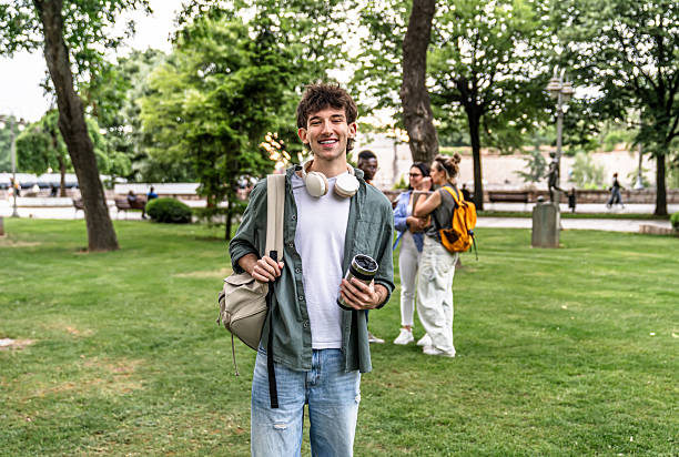 Young male student smiling at camera while walking in a park with his friends in the background