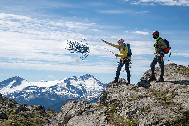 Climbing guide throws rope over cliff as guest watches, on mountain top, British Columbia Coastal Ranges