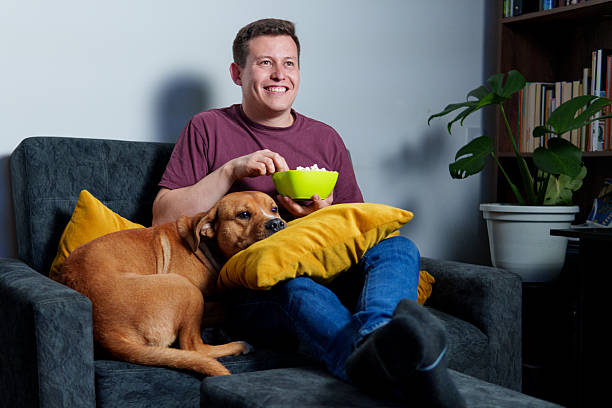 istock Man eating popcorn watching tv with dog on sofa at home 2225139418