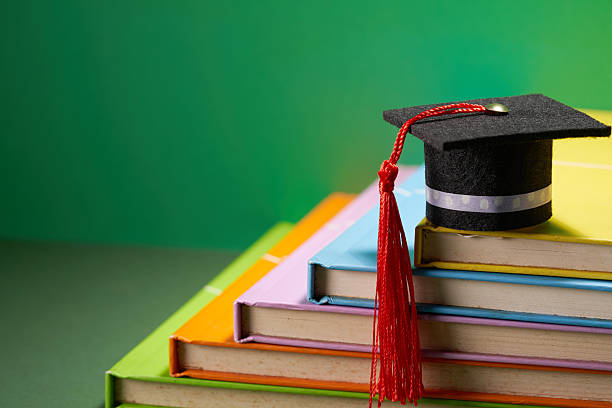 Mortar board and stack of books on a desk in front of chalkboard.