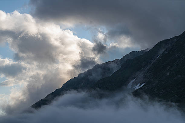 A majestic mountain landscape features dark rocky peaks, enveloped by low-lying fog and illuminated by dramatic clouds, evoking a moody yet captivating ambiance.