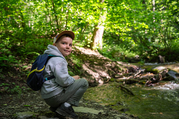 A cheerful young boy wearing a backpack enjoys exploring the forest and a nearby stream, engaging with the natural surroundings under bright daylight, embracing outdoor adventure.