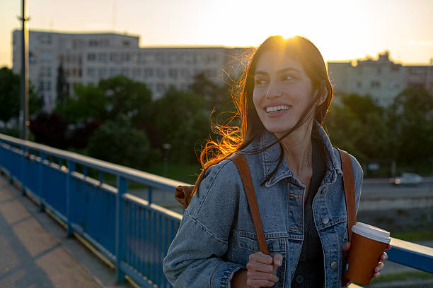 istock Young Woman Smiling and Holding Coffee While Walking on Bridge at Sunset 2225152906