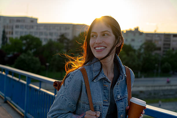 istock Young woman smiling while walking on a bridge at sunset with takeaway coffee 2225152912