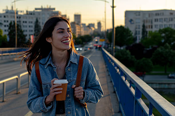 istock Young woman smiling while walking on a bridge at sunset, drinking coffee 2225152989