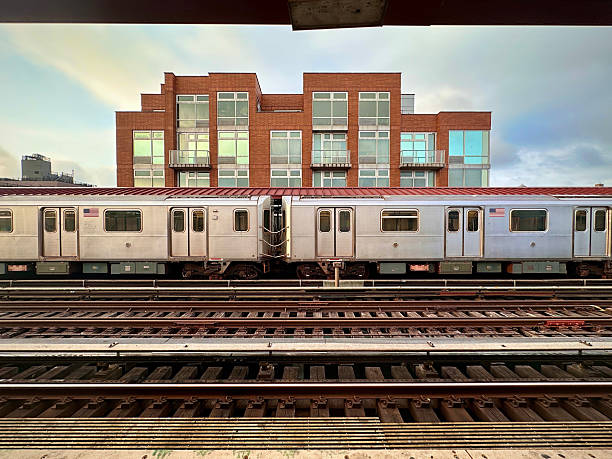 A stationary subway train sits in front of a modern red-brick apartment building, with multiple glass balconies reflecting the soft light of early evening.