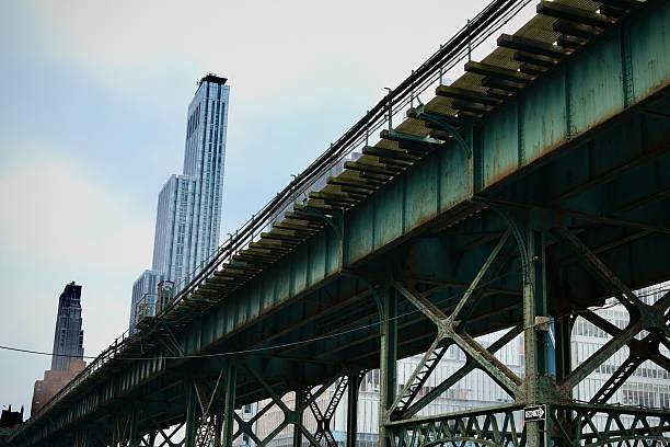 A rusting green train bridge angles sharply through the image as glass skyscrapers rise in the distance, evoking tension between industrial infrastructure and modern vertical growth.