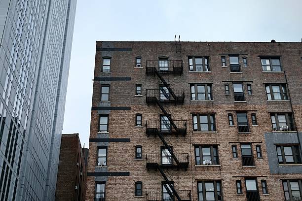A tightly gridded brick apartment facade is lined with black fire escapes, pressed up against the looming wall of a neighboring glass tower.