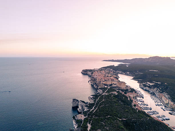 Aerial view of Bonifacio town perched on limestone cliffs with marina and boats at sunset. Scenic coastal landscape with rugged cliffs, Mediterranean Sea, and hillside greenery in Corsica. Coastal town and harbor panorama.