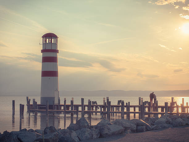 A tall red and white striped lighthouse stands by a calm lake at sunset. A couple is silhouetted near the dock, enjoying the serene atmosphere and beautiful twilight.