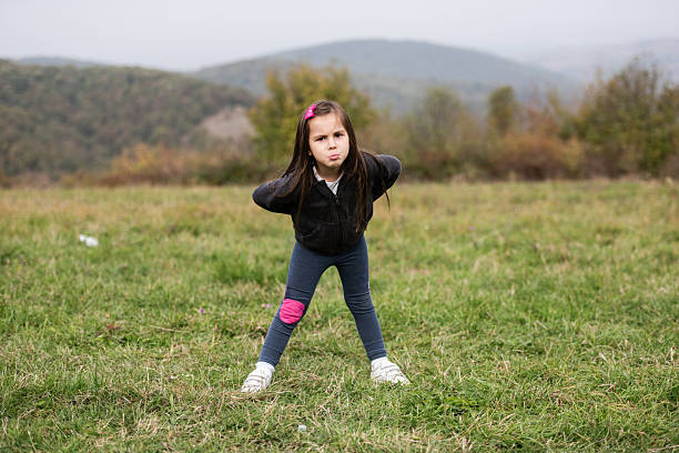 Young girl making a funny face while standing with legs apart and hands on hips in a grassy meadow with hills in the background