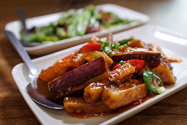 Close-up of stir-fried eggplant with chili sauce and sliced peppers served on a white plate at a restaurant table.