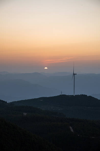 The drone rises with the first light, revealing rows of white turbines perched on a forested ridge. Low-lying fog pools in the valleys, catching pink and amber rays that spill across the turbine towers and spinning blades.Their tips glowing where sunlight kisses the treetops. The motion of the rotors is slow and steady, synchronized with the gentle climb of the camera, conveying both power and serenity. This clip is ideal for stock libraries covering renewable energy, aerial cinematography, sunrise landscapes, mountain scenery, and sustainable-development storytelling.