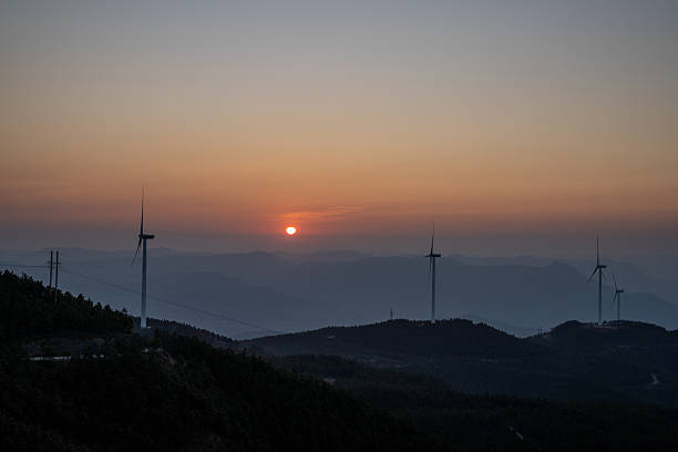 The drone rises with the first light, revealing rows of white turbines perched on a forested ridge. Low-lying fog pools in the valleys, catching pink and amber rays that spill across the turbine towers and spinning blades.Their tips glowing where sunlight kisses the treetops. The motion of the rotors is slow and steady, synchronized with the gentle climb of the camera, conveying both power and serenity. This clip is ideal for stock libraries covering renewable energy, aerial cinematography, sunrise landscapes, mountain scenery, and sustainable-development storytelling.