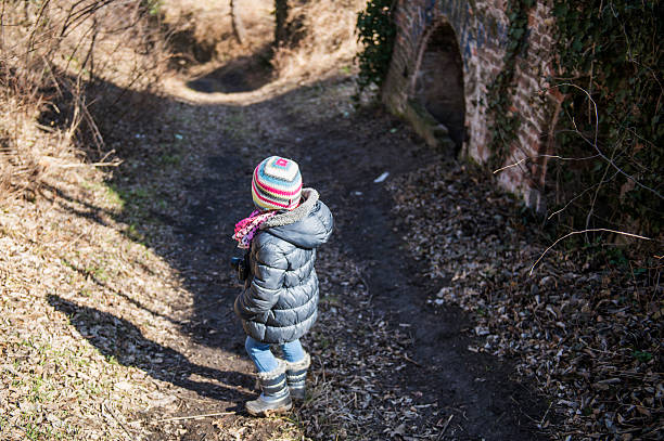 Young girl walking on a path near an old brick wall in nature, holding a camera and wearing winter clothes
