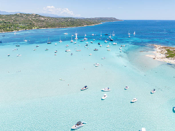 Numerous boats anchored in shallow, crystal-clear turquoise water near a sandy beach and green hills. Scenic coastal bay with sailboats and motorboats. Tropical seascape with leisure vessels.