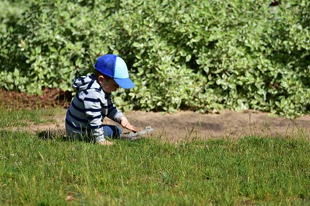 Young boy playing with plastic container in sandpit on sunny day. Child wearing striped hoodie and blue cap enjoying imaginative outdoor play, symbolizing curiosity, learning, and childhood fun.