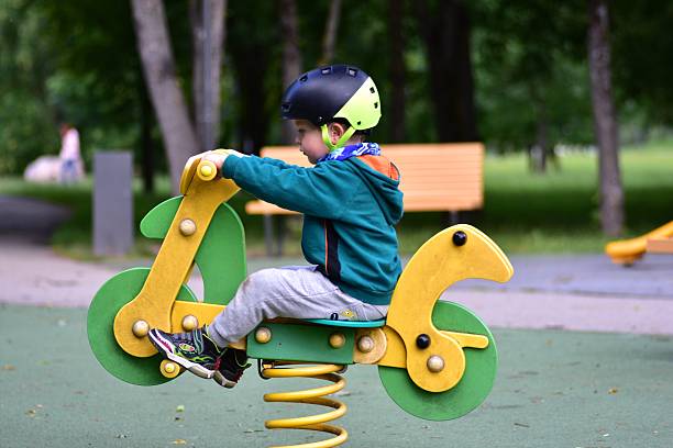 Boy bouncing on spring rocker shaped like a motorcycle in playground, wearing helmet and green hoodie. Joyful scene captures movement, excitement, and childhood playfulness in colorful outdoor space.