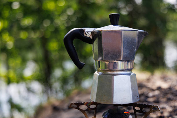 A silver moka pot sits on a camping stove, surrounded by a forest backdrop.