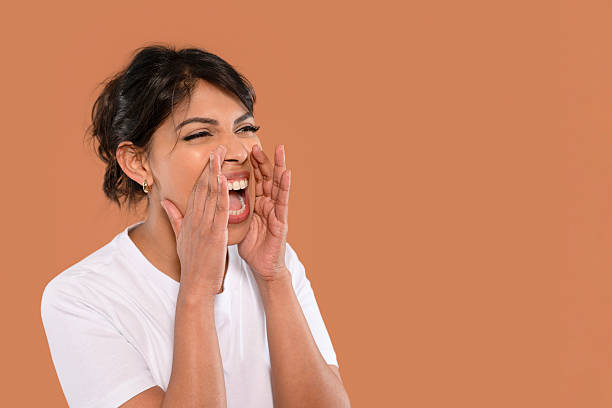 A woman is joyfully shouting with her hands cupping her face, radiating enthusiasm and positivity, highlighting a vibrant and lively moment against a warm backdrop.