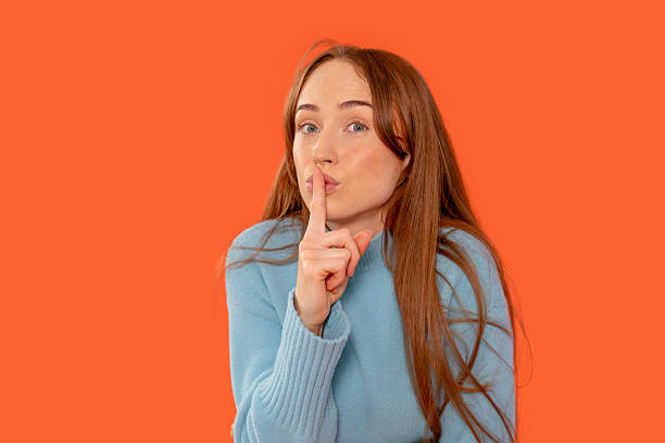 Woman gestures for silence with finger on lips against bright orange background, showcasing playful expression and serene demeanor during a quiet moment