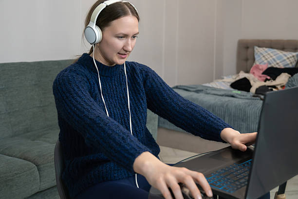 istock Young woman wearing headphones is working or studying from home, using laptop and mouse, concentrating on the screen while sitting in her messy bedroom 2225198006