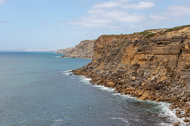 Dramatic Atlantic coastline near Azenhas do Mar, Portugal, featuring rugged cliffs, ocean waves, and layered rock formations. A cloudy sky casts soft light over the expansive coastal landscape, showing the natural beauty and geological detail of the region.