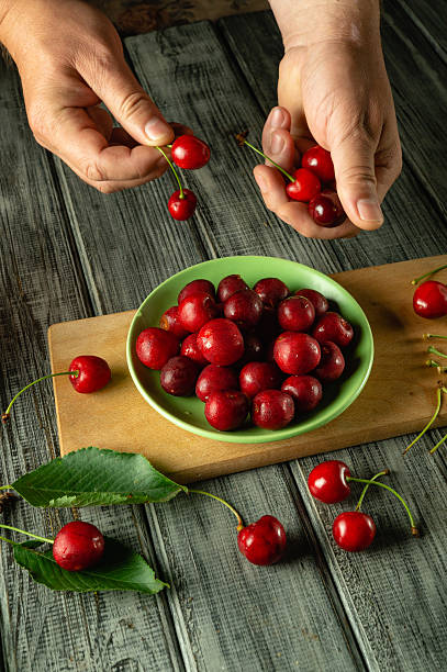 istock Hands carefully select and sort ripe cherries on a wooden table. A green bowl holds a bountiful harvest of red cherries, showcasing nature's bounty and seasonal delight 2225202002