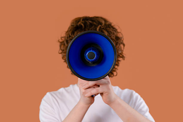 istock Person holds blue megaphone against a plain orange background while engaging in lively communication to express ideas and gather attention from an audience 2225203097