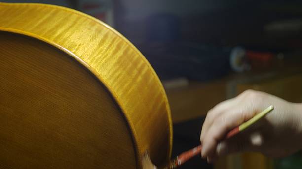 istock Skilled luthier carefully brushing deep-amber varnish across polished cello wood, sunlight illuminating intricate handcrafting process in traditional workshop setting 2225207647