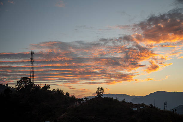 A picturesque hillside in an Indian village of Uttarakhand glows under a vibrant sunset. A tall cell tower rises among trees, blending modern connectivity with serene natural beauty.