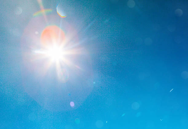 A brilliant sun creates powerful light rays and lens flares against a deep blue sky, representing the dangerous intensity of summer heatwave conditions. The stark sunburst effect and bright solar radiation illustrate the extreme heat and UV exposure that threatens public health during scorching weather events.
