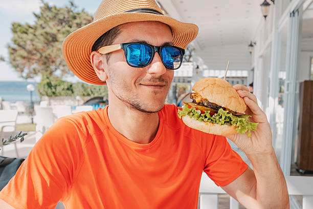 Young man wearing sunglasses and straw hat holding a burger in outdoor restaurant with sea view