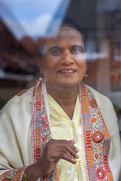 Close-up portrait looking through a window of a senior adult female wearing traditional clothing, smiling at the camera while standing in a home in Newcastle-upon-Tyne, North East England.Videos similar to this scenario available.