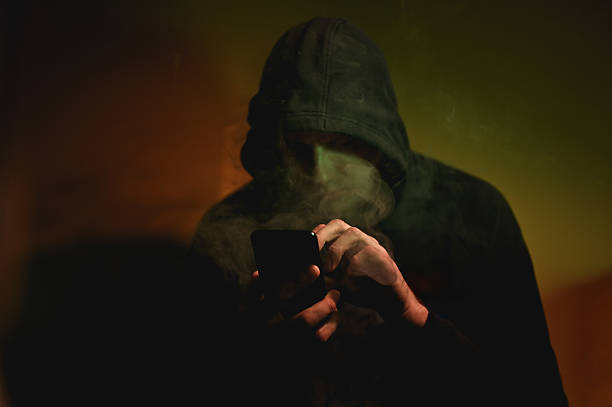 istock Hooded man smoking and using smartphone in dark room 2225213966