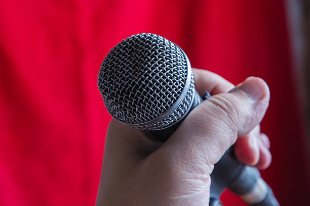 istock Hand holding a microphone on a microphone stand against a red curtain background close-up, dynamic microphone for singing with hand, karaoke, podcast, and live broadcast, singing performance. 2225227976