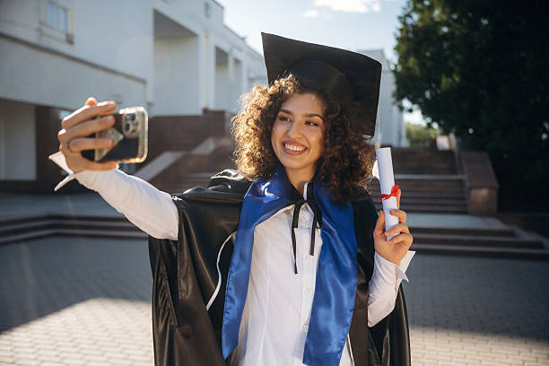 istock Making a selfie, holding diploma document. Cheerful graduate student in a gown is standing outdoors 2225228124