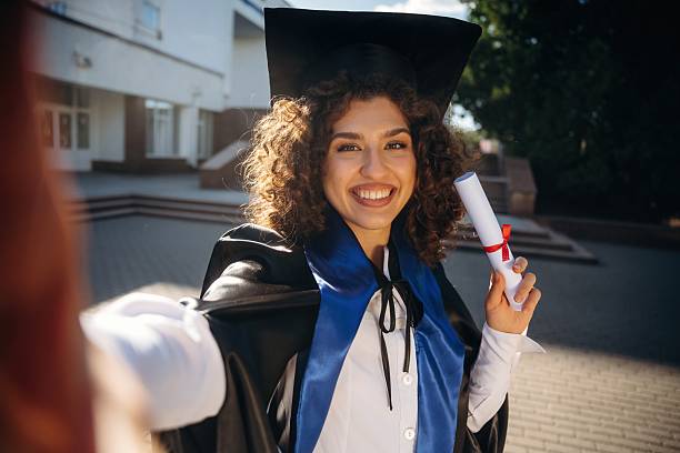 istock Making a selfie, holding diploma document. Cheerful graduate student in a gown is standing outdoors 2225228129