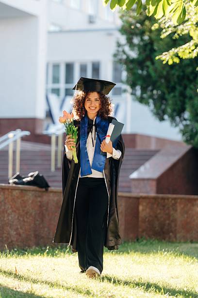 istock Green tree and university building behind. Cheerful graduate student in a gown is standing outdoors 2225230010