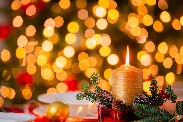 Red Christmas burning candle on cozy dining table illuminated by defocused glittering lights at background. Greeting card or copy space. Holiday or New Year's Eve decoration
