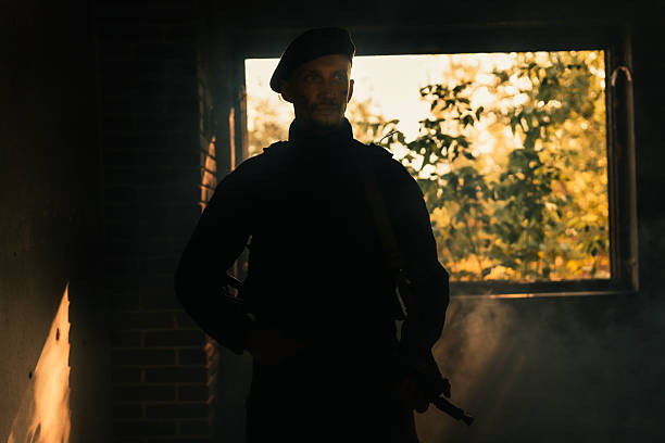 Silhouetted figure of an armed individual standing in a dimly lit room with sunlight streaming through a window, conveying themes of vigilance, security, and isolation.