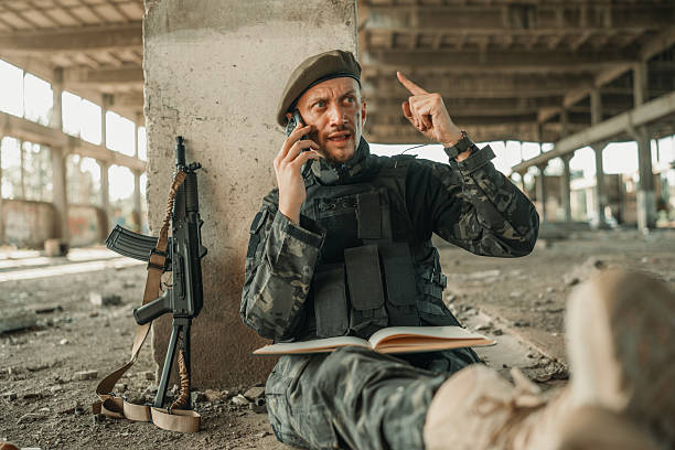 A soldier sits in an urban setting with tactical gear, holding a phone for communication, surrounded by a firearm and notepad in an abandoned building.
