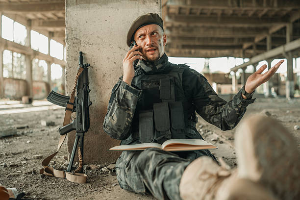 A military soldier in tactical gear makes a call using a cellphone while sitting in an empty industrial building. A modern weapon leans on the wall beside him, conveying themes of military operations and technology.