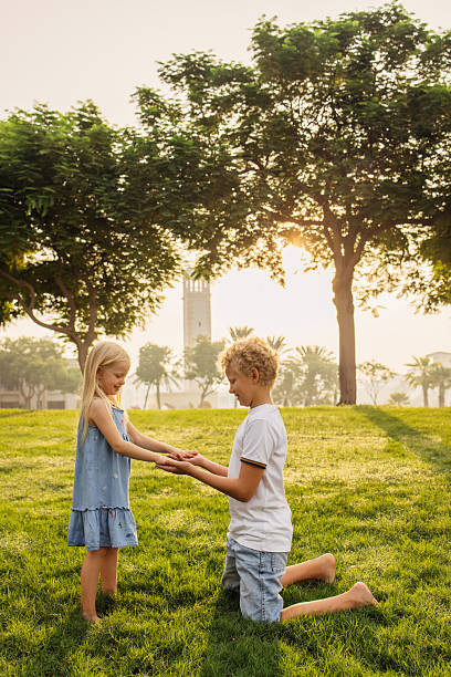 A harmonious scene in a lush park during sunlight. A boy and a girl interact playfully, conveying happiness, togetherness, and youthful moments. The backdrop is filled with greenery and an architectural landmark towering at the horizon, creating a peaceful mood of joy and childhood simplicity.